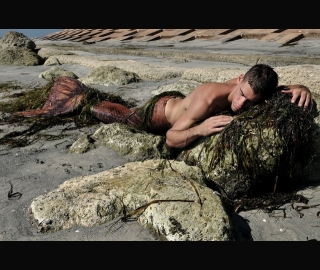 Merman laying on rocky beach strewn with seaweed